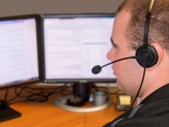 A technical support assistant sits in front of two computer monitors wearing a headset with microphone.